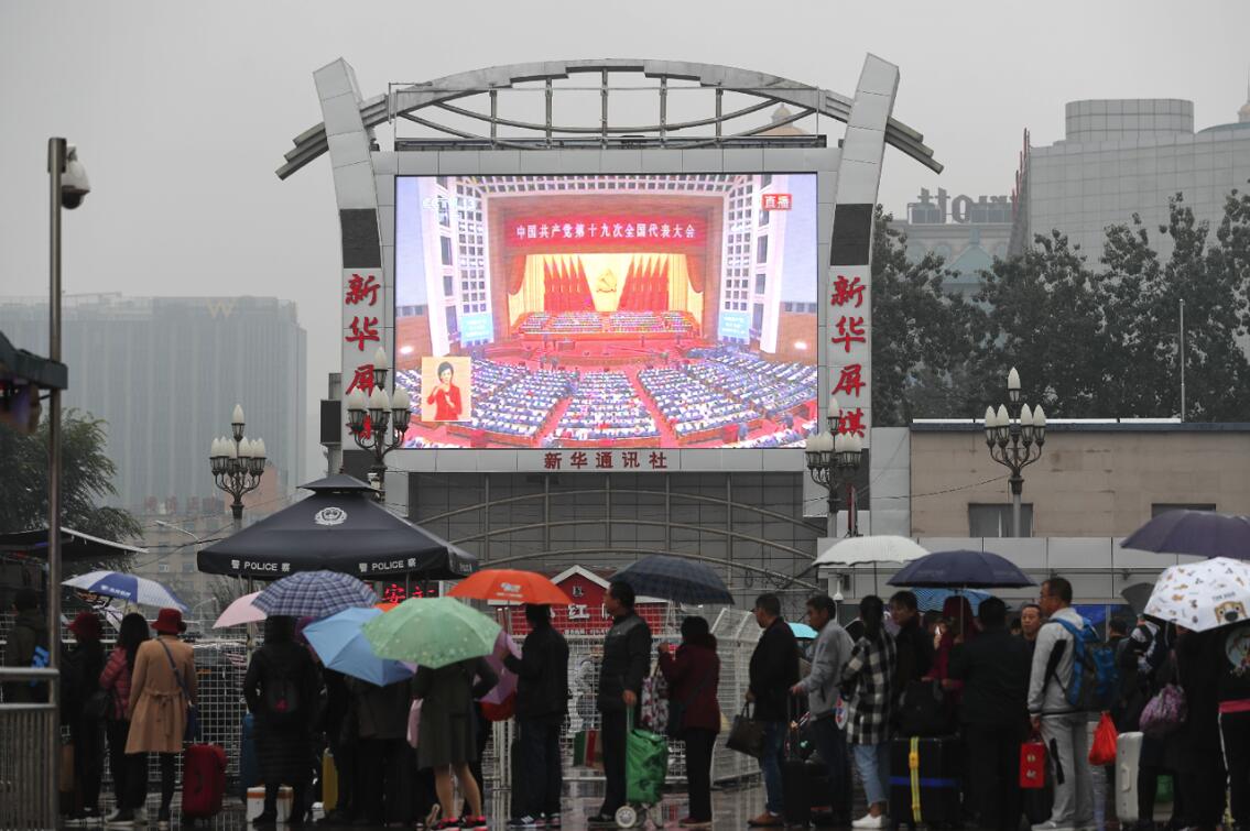 北京火车站站前，民众在雨中驻足观看十九大开幕式直播